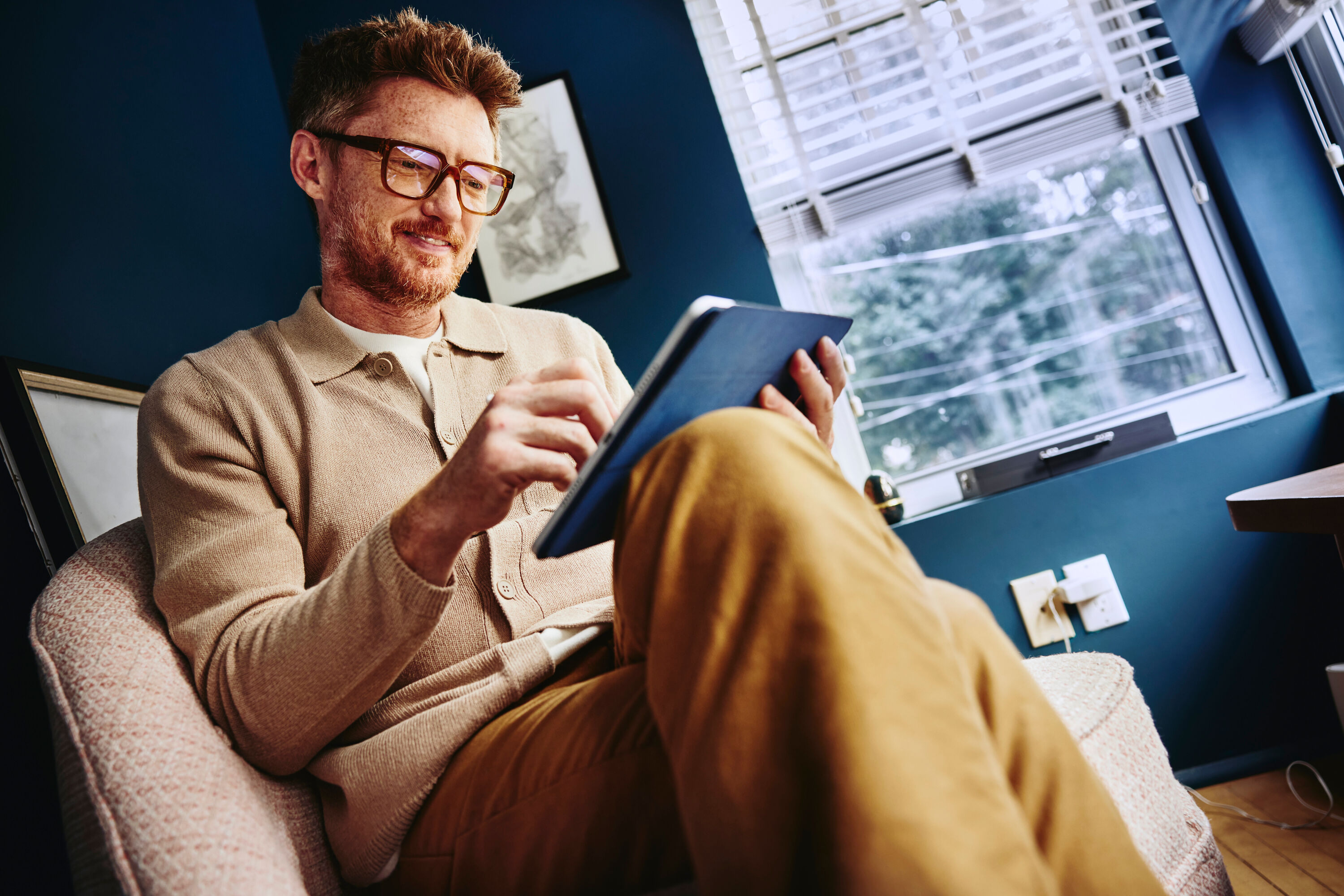 man looking at tablet wearing glasses, legs crossed, wearing brown clothing