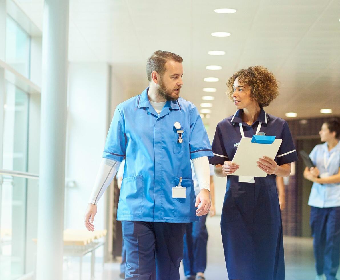 Male and female healthcare professional walking together in a hospital setting. The female is carrying a clipboard.