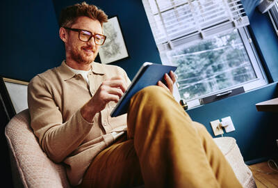 man looking at tablet wearing glasses, legs crossed, wearing brown clothing
